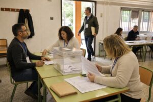 Interventor sentado junto a una mesa electoral y apoderado de pie en la entrada de un local de votación